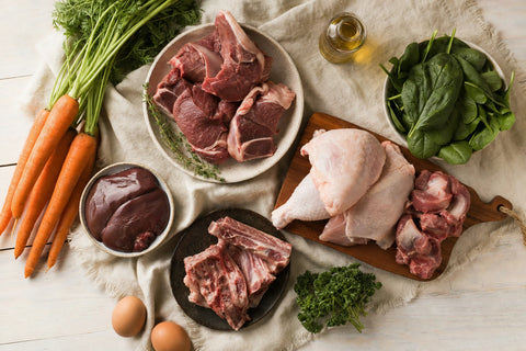 Raw dog food ingredients including meat, offal, and vegetables laid out on a linen surface